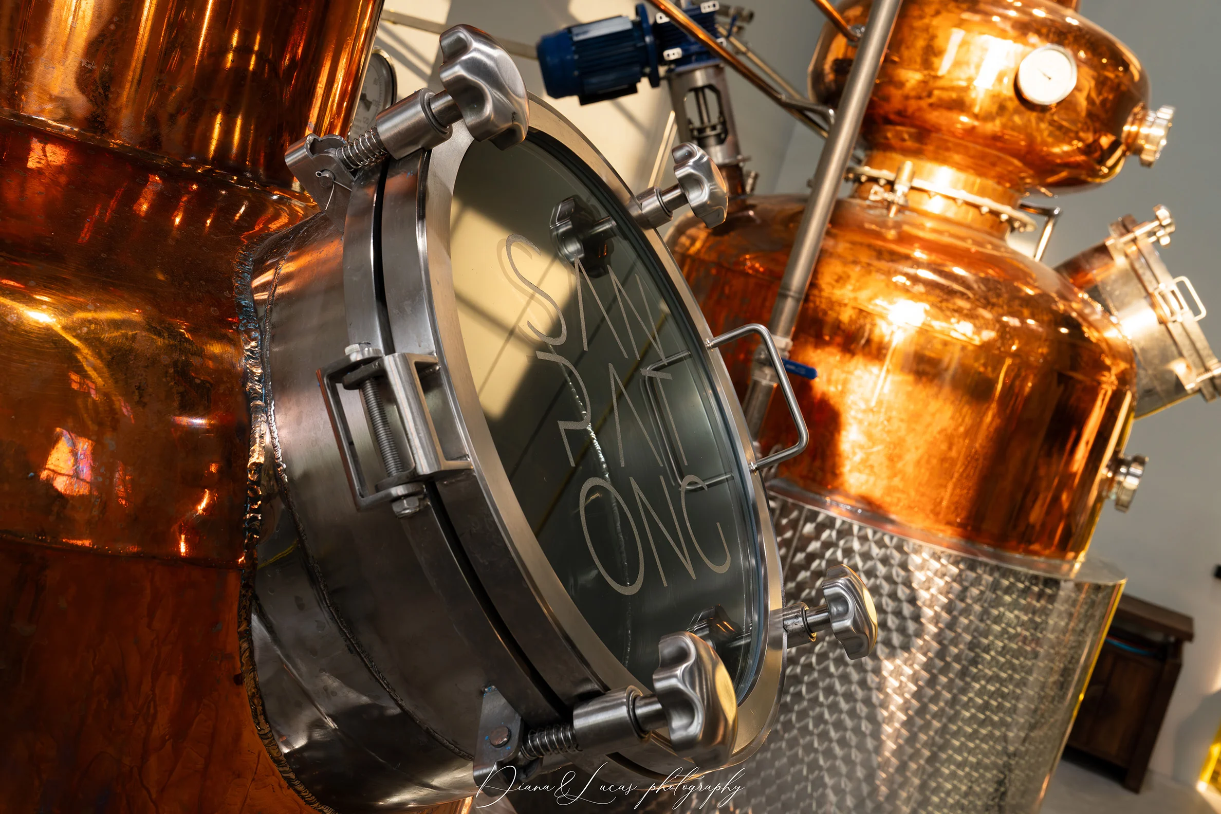 Close-up of copper distillation equipment with a round glass inspection window, stainless fittings, and warm reflections in an industrial setting.