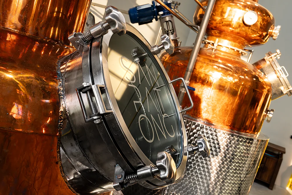 Close-up of polished copper distillery stills with metal manway, pipes, valves, and hammered steel tank in a spirits production facility